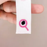 Symbole œil brodé en rose sur le bas du ruban du marque-page Grizzle – détail couture